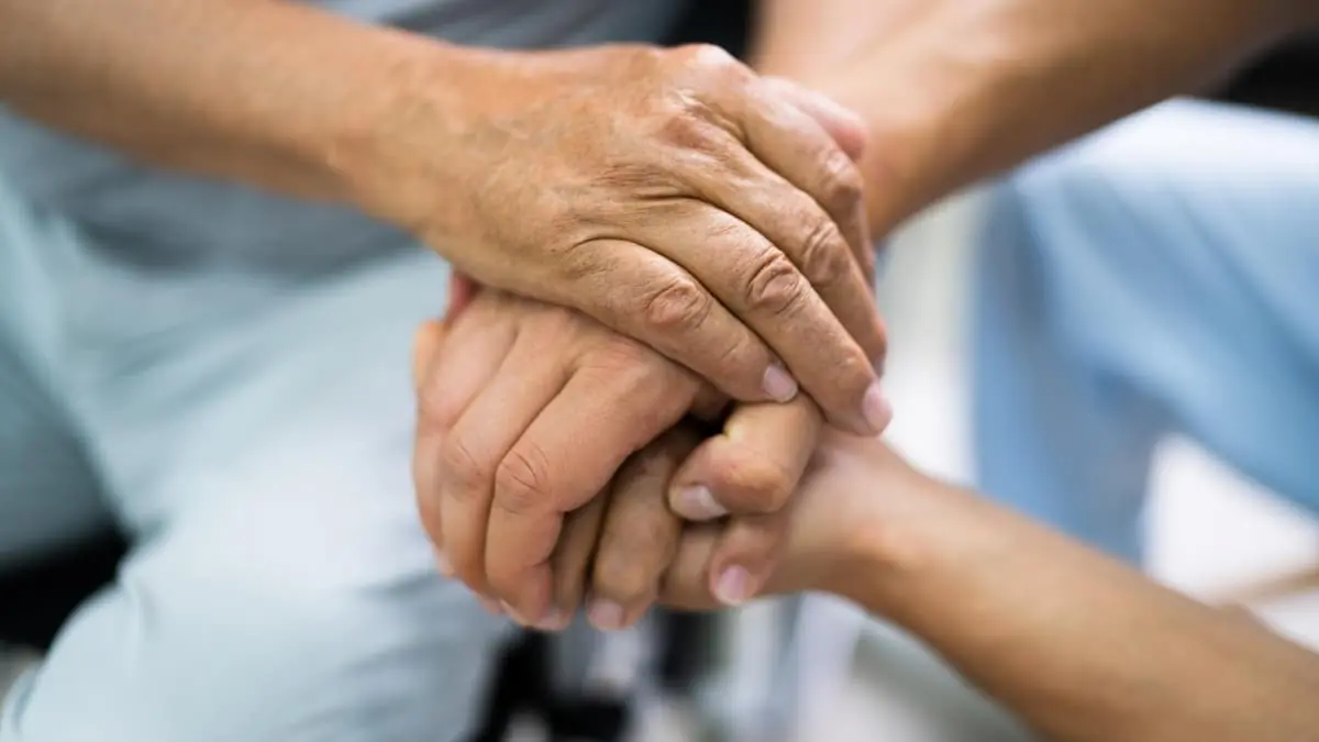 Close-up of an older person’s hands held gently between a caregiver’s hands