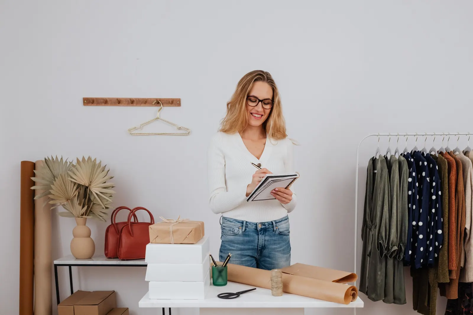 A shop owner stands in her bright new boutique, making notes on a clipboard beside a rack of clothing and wrapped parcels.