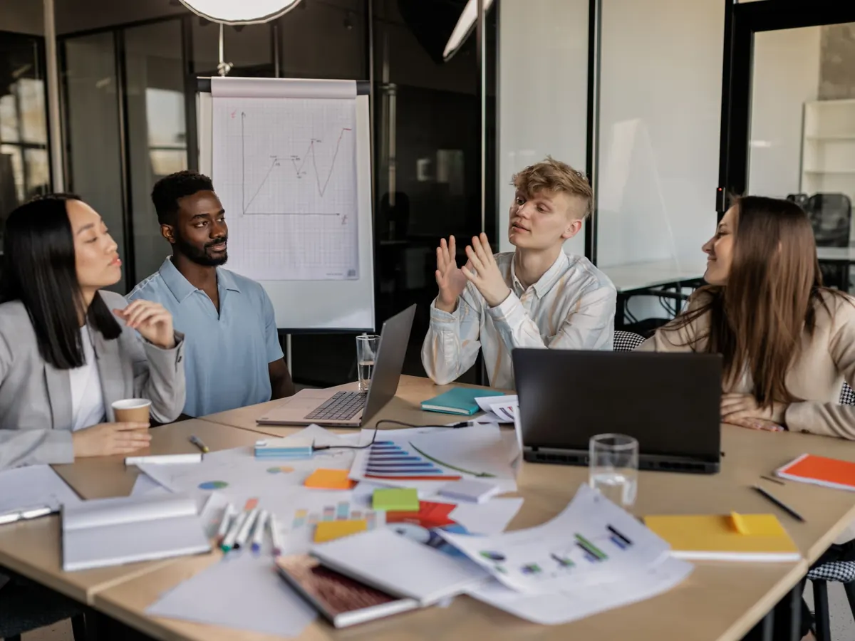 A small team of four talks around a meeting table covered with notes, charts and laptops.