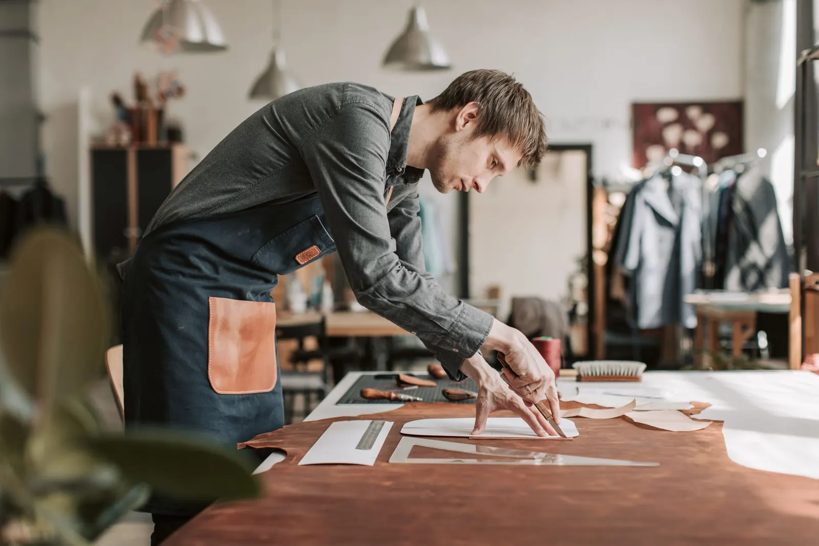 A leatherworker in an apron leans over his workbench, cutting leather pieces in his own sunlit shop.
