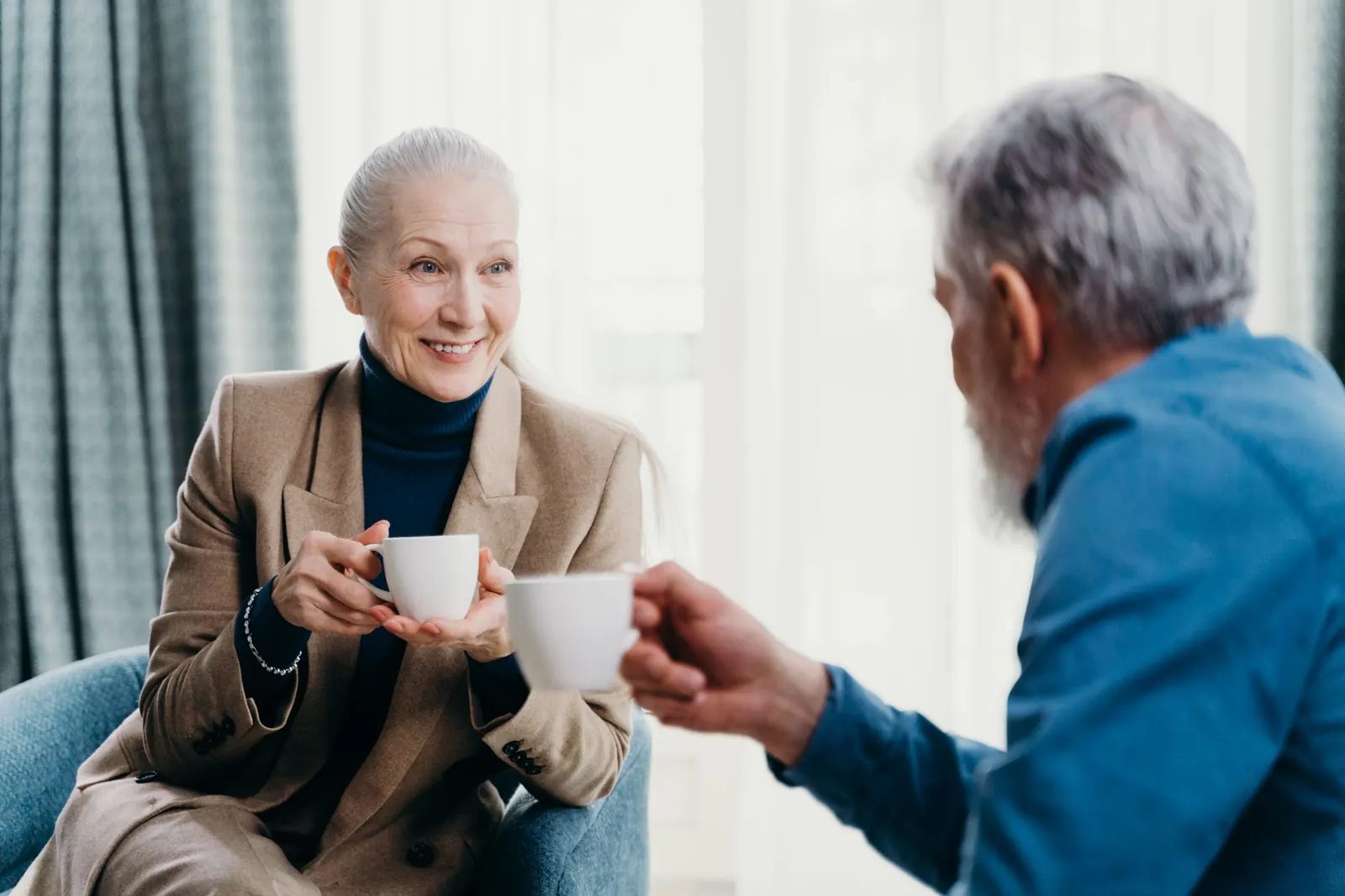 An older couple shares coffee in a sunlit living room, the woman smiling as she talks with her husband.