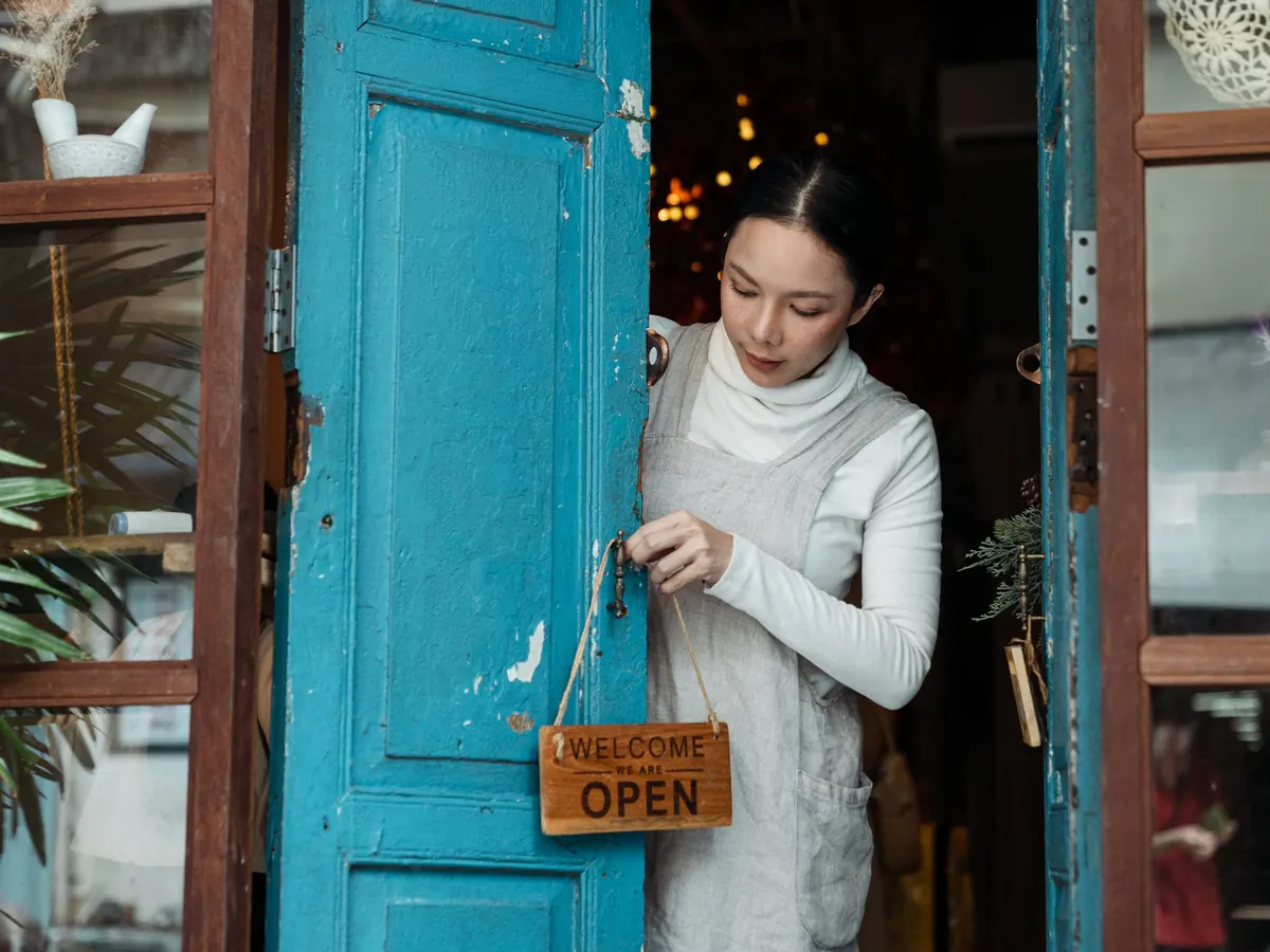 A shop owner in an apron hangs a wooden Welcome We Are Open sign on the handle of a blue door.