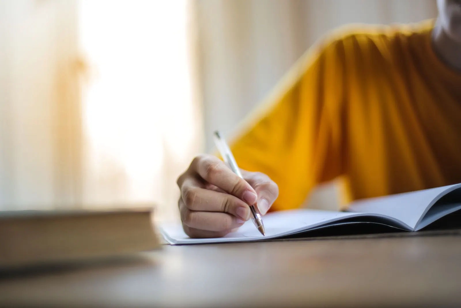 A hand writes in an open notebook at a table beside a bright window.