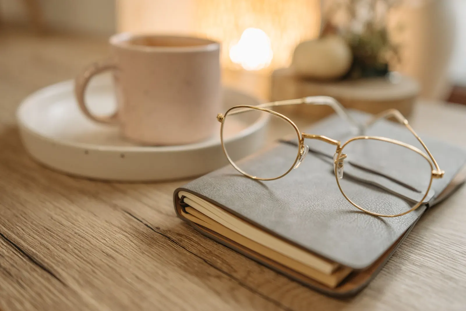 Gold rimmed reading glasses rest on a leather notebook beside a ceramic coffee cup on a wooden table in warm light.