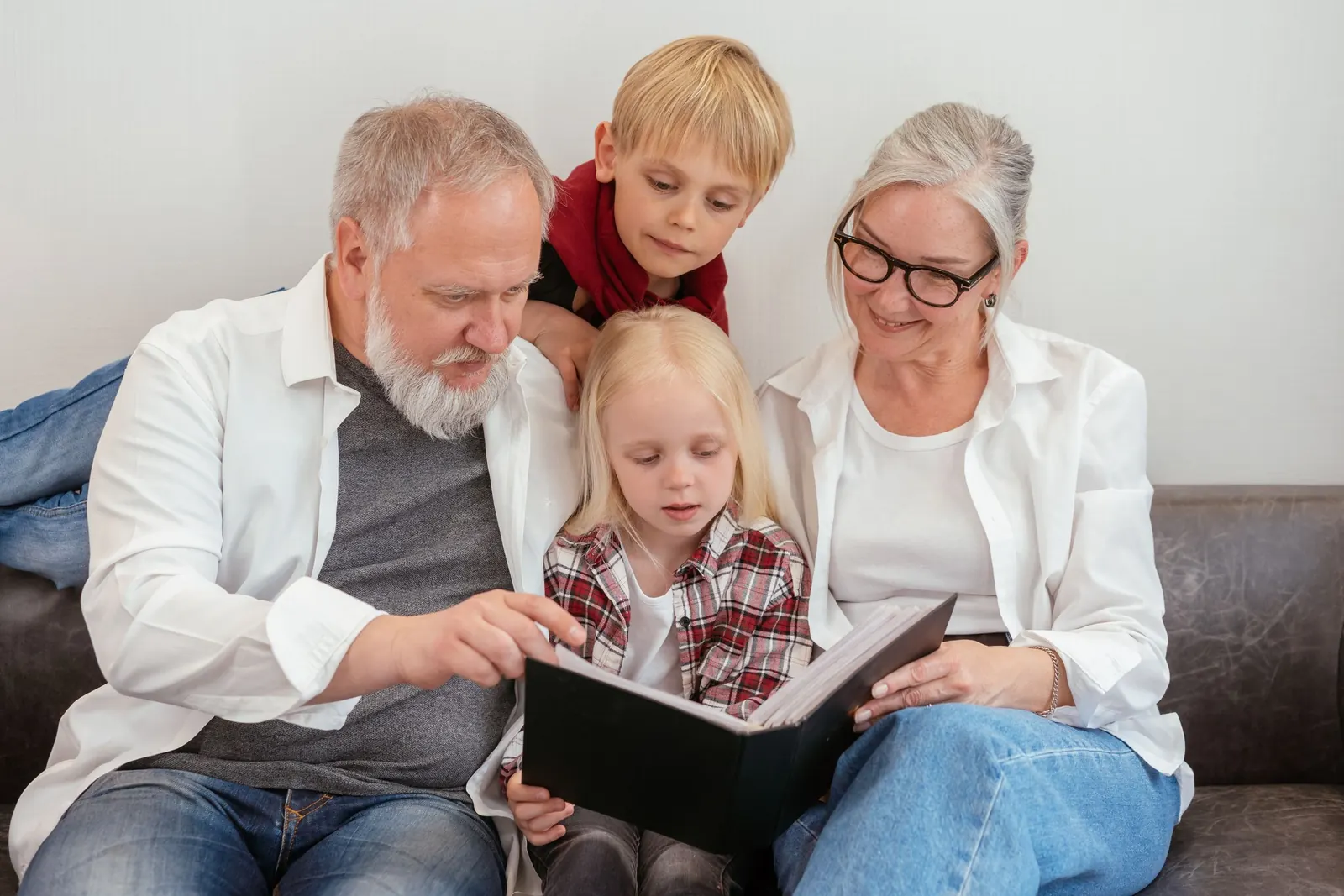 Grandparents on a sofa read a book with their two young grandchildren, one leaning over the grandfather's shoulder.