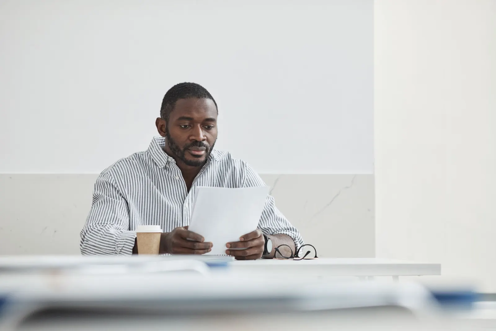 A man in a striped shirt sits at a white desk reading through a printed document, a coffee cup at his side.