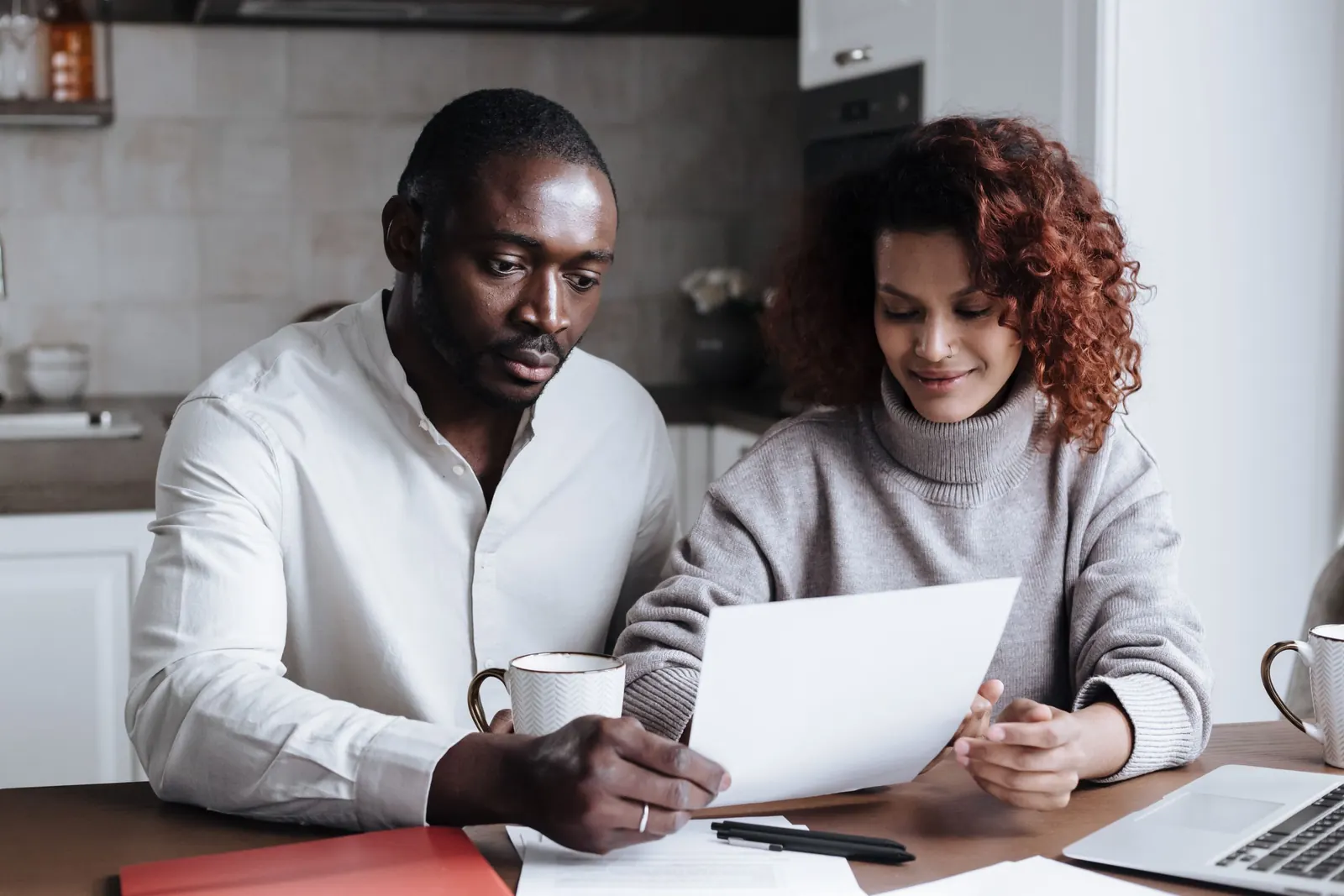 A couple sits at their kitchen table reading a printed document together, a coffee mug and laptop beside them.