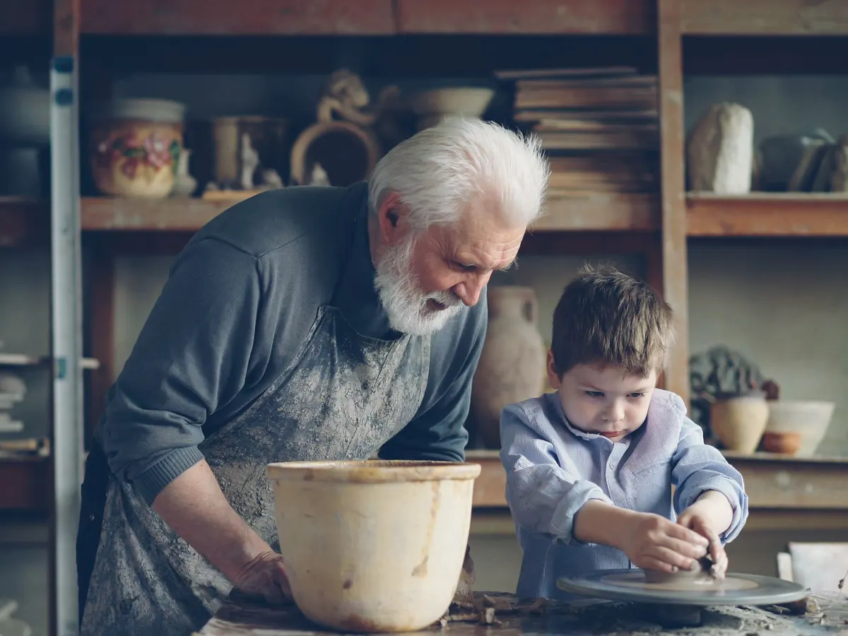 A white bearded potter leans over his wheel while his young grandson shapes clay beside him in a workshop lined with pottery.