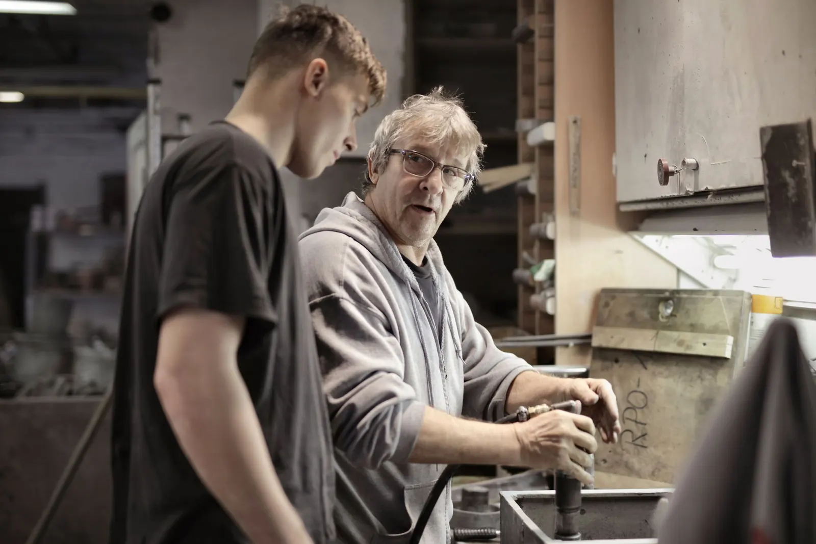 An older craftsman in glasses works at the bench while a younger man stands beside him, watching closely in a workshop.