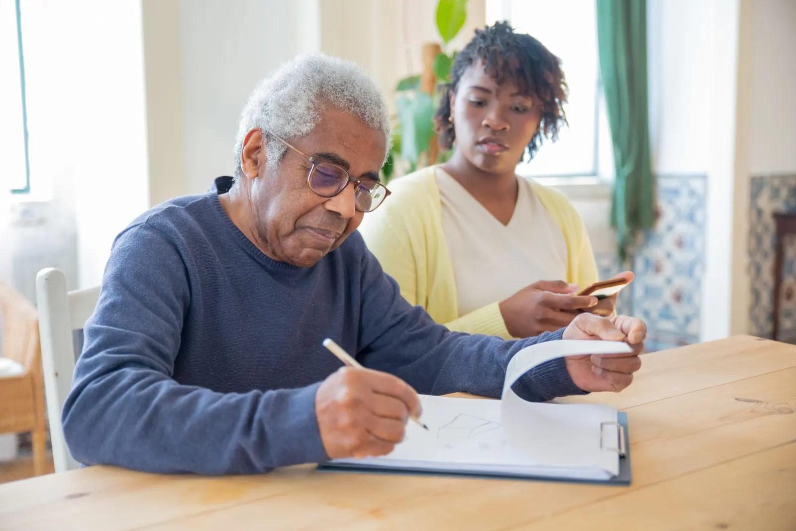 An older man writes on a clipboard at a wooden dining table while his adult daughter sits beside him, following along.